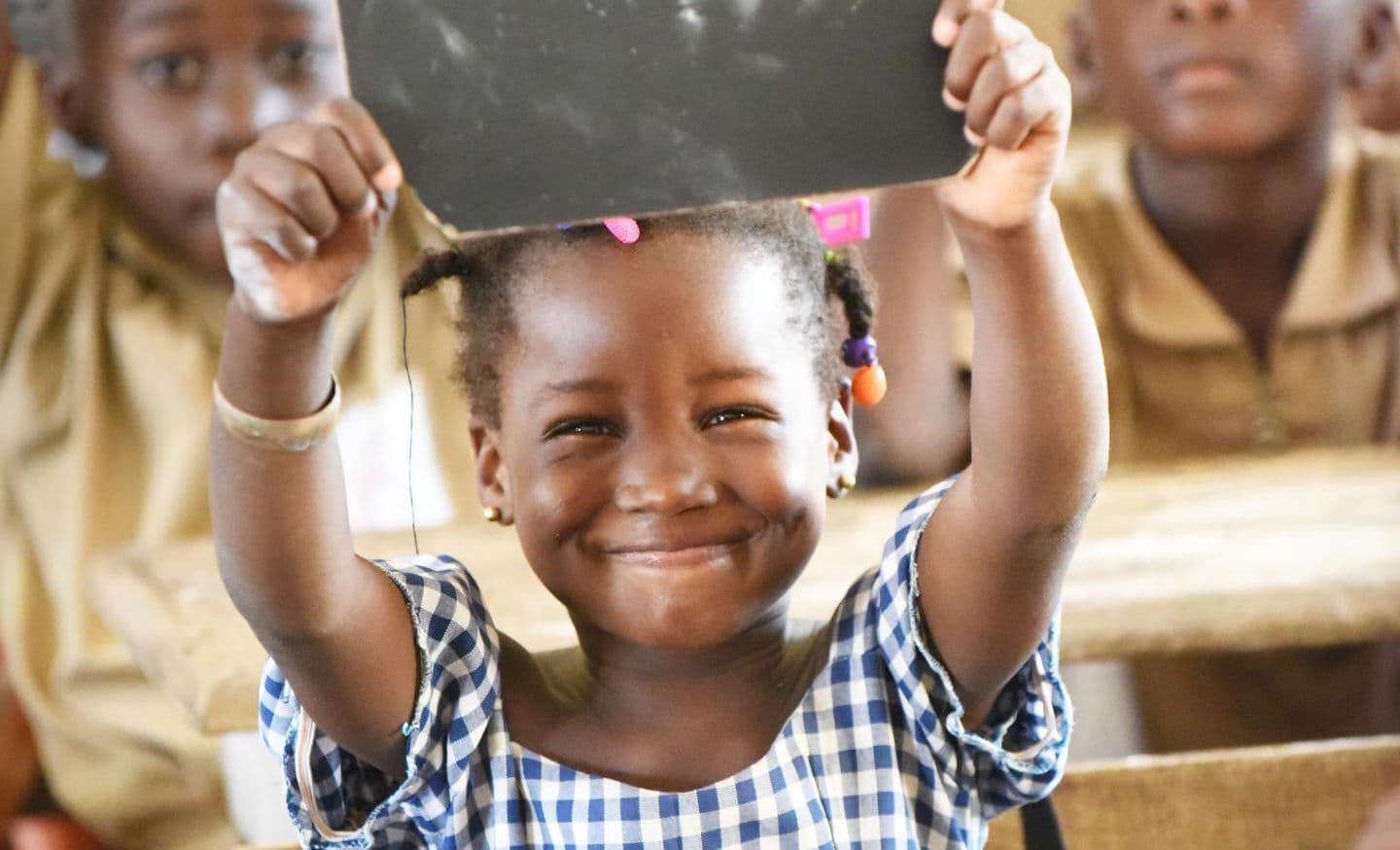 A smiling girl in a classroom holds a chalk slate above her head
